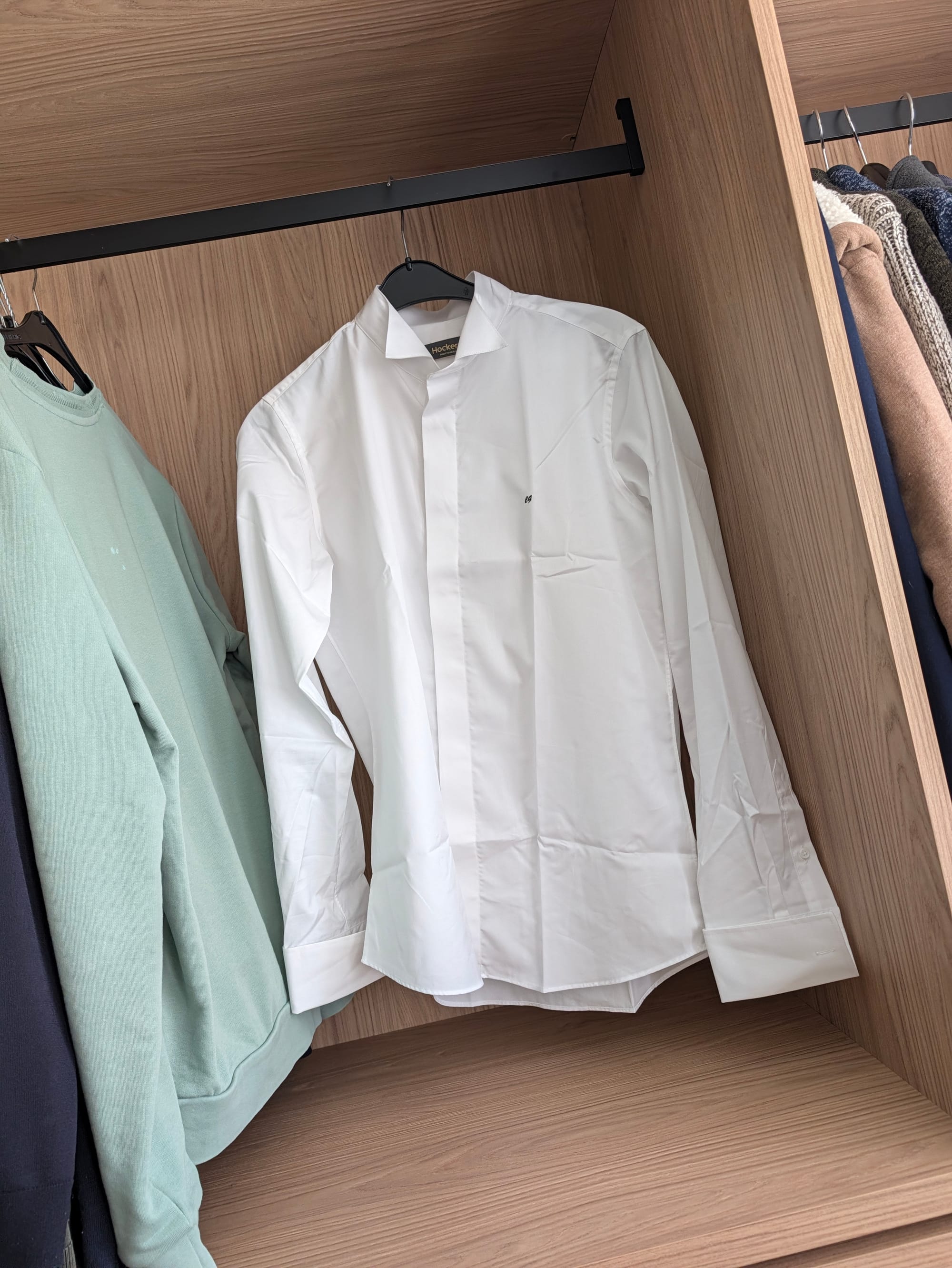 Chemise blanche sur mesure Hockerty avec col cutaway et logo brodé, suspendue dans un dressing