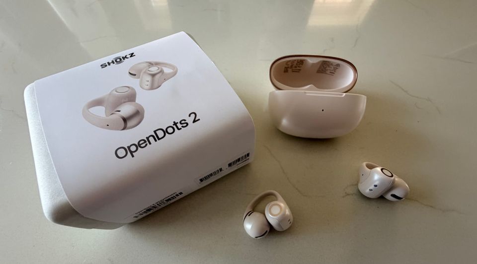 Shokz OpenDots 2 en pearl white posés à côté de leur boîte et du boîtier de charge ouvert sur un plan de travail