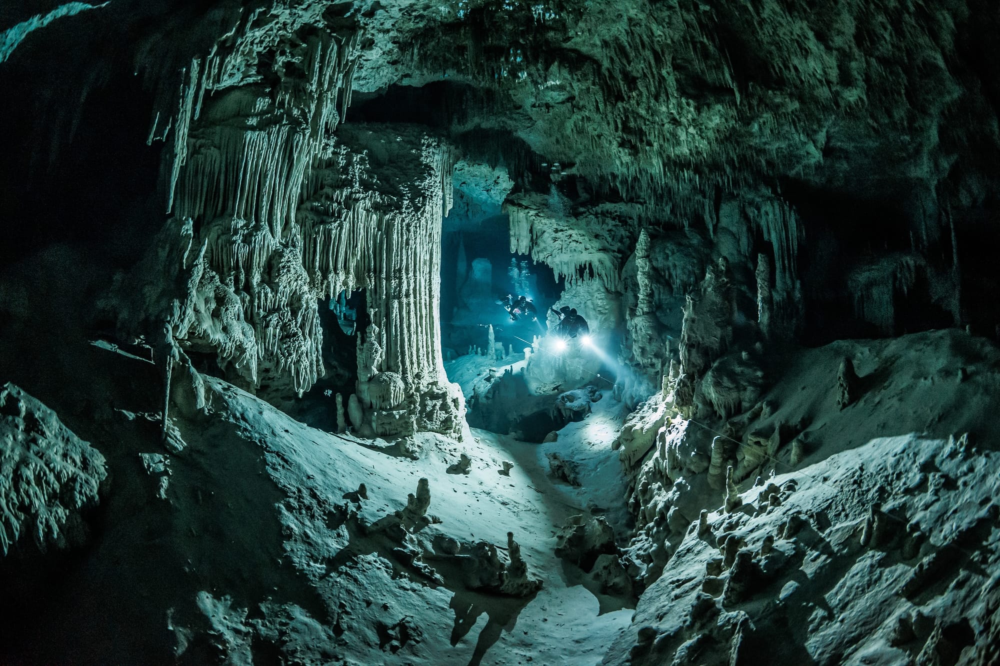 A wide, grey, rocky, underwater, water filled cave, with cave divers inside