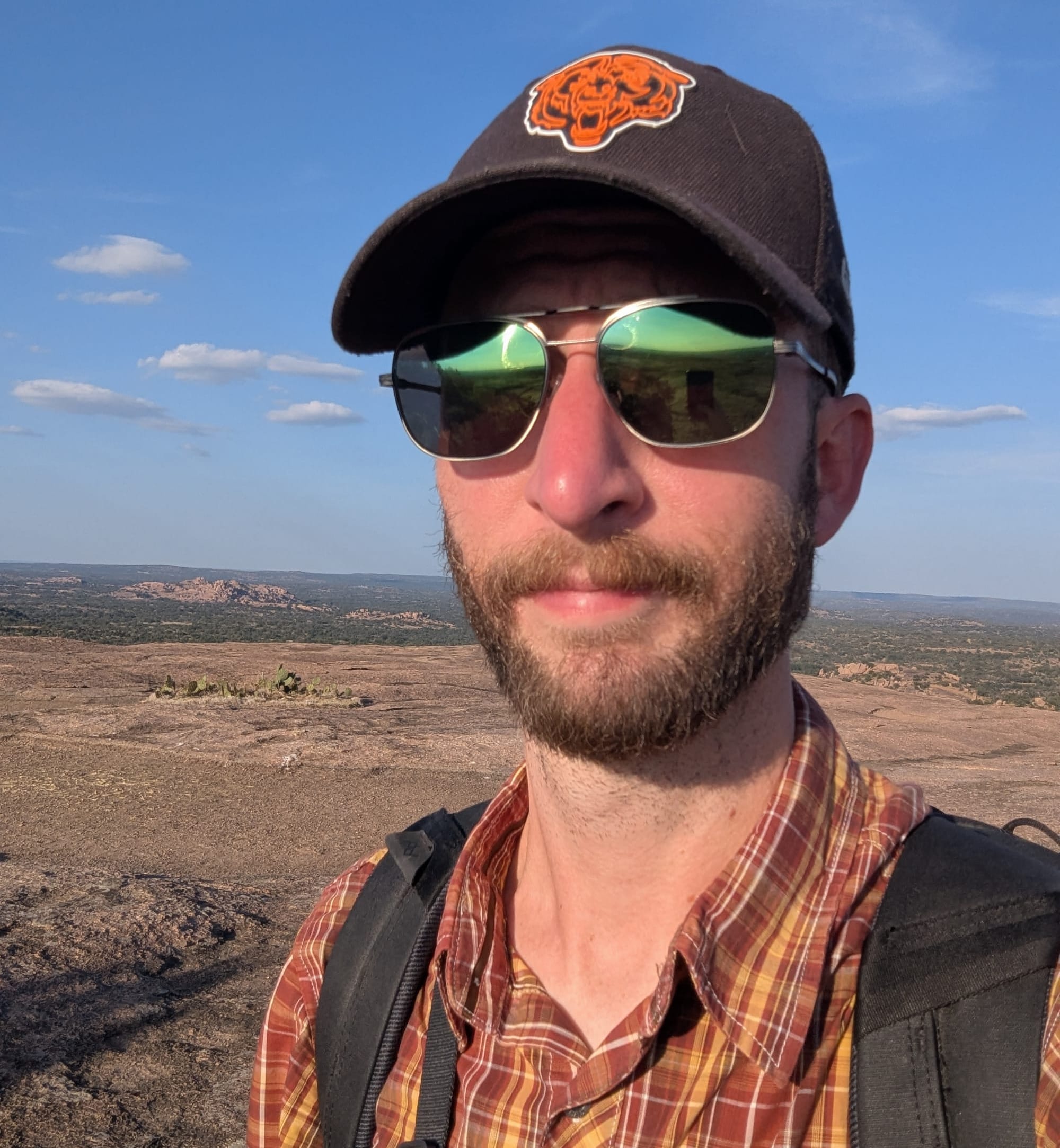 Selfie photo of a man in a hat and sunglasses with a backpack, standing on bare rock with a view of isolated rocky hills on the horizon.
