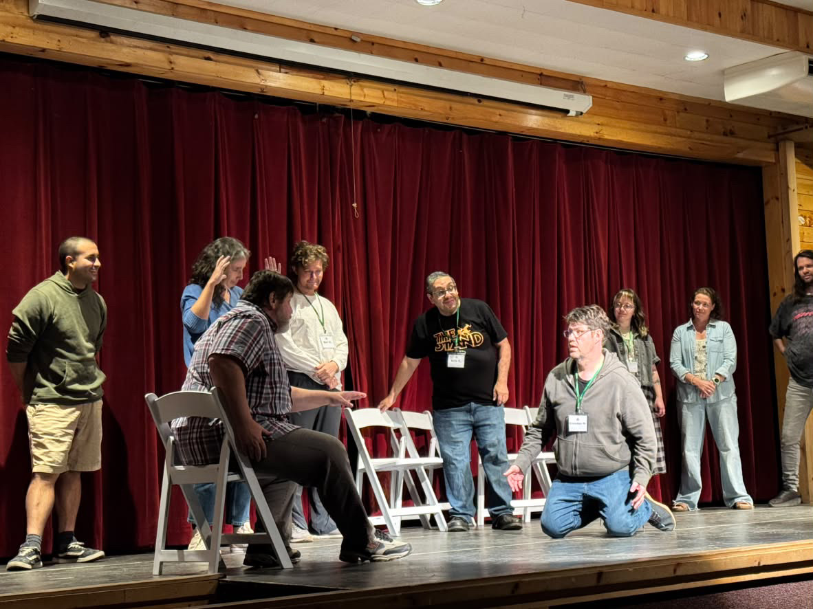 Picture of improvisors on stage. Seven stand in a backline in the back while two others are in the fore ground. One is seated, the other kneels.