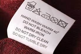 A close-up of a clothing care label with instructions for washing and care. It includes symbols for hand washing, avoiding bleach, warm ironing, not dry cleaning, and not tumble drying