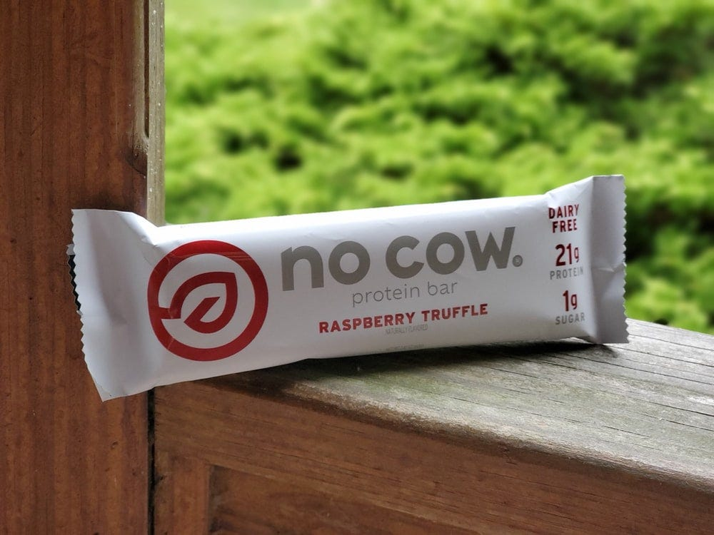 This image shows a “No Cow” protein bar in raspberry truffle flavor, emphasizing its high protein content (21g) and low sugar (1g) with the label “dairy-free.” The bar is designed to be a nut