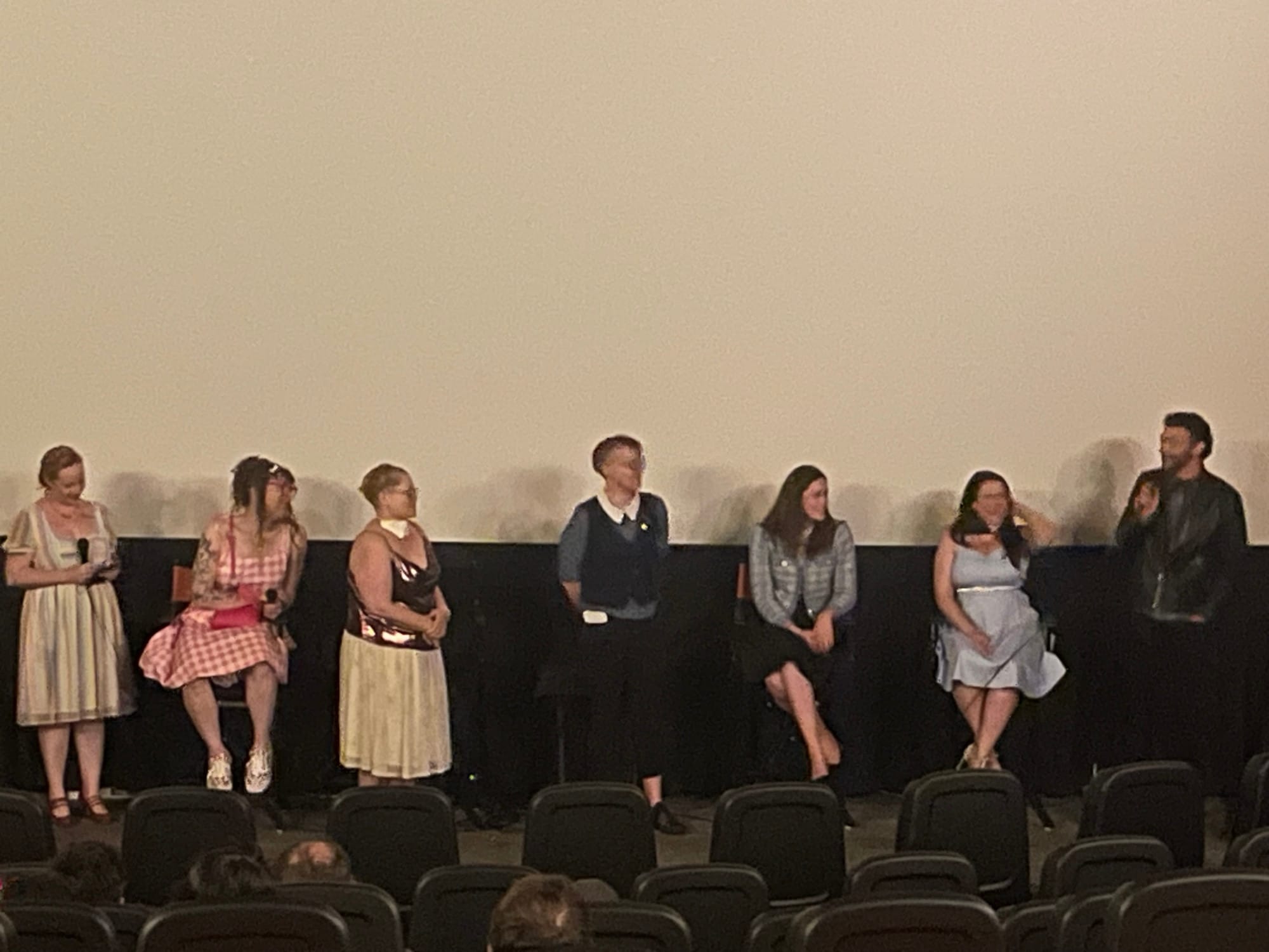 A shot of Long Away cast and crew doing a panel discussion in a movie theater, in front of the screen, featuring producer Dr. Erin Macdonald, Tilly, Susan, Jessie Earl, Griffin Kelly, Jewel Cavazos, and Justin Lawrence Barnes