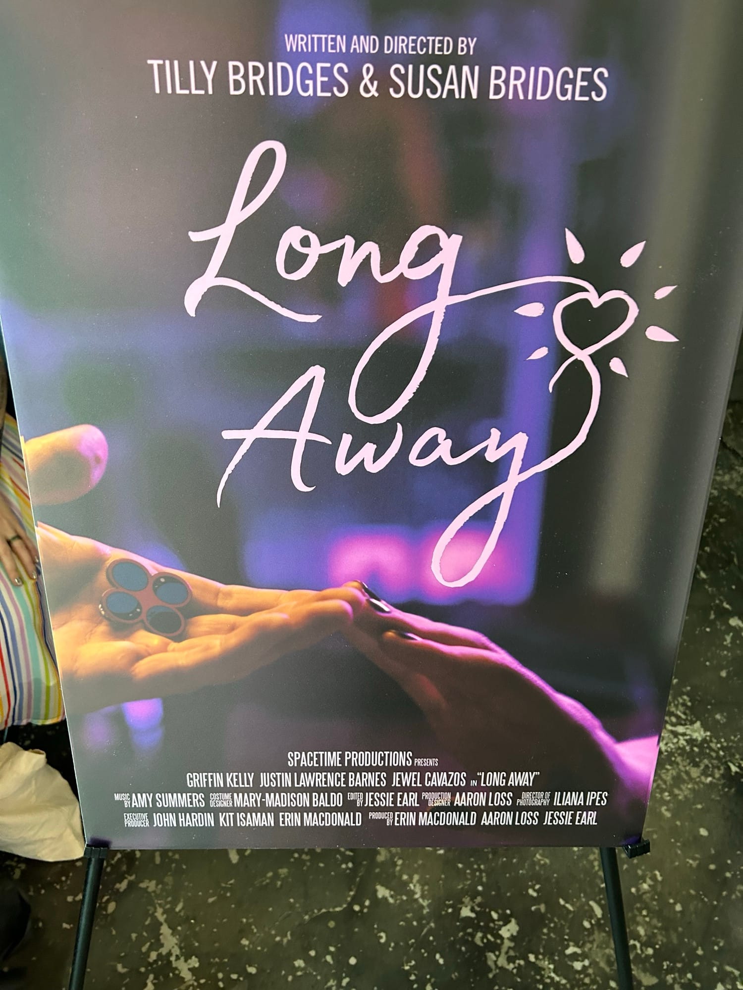 one hand reaching out, palm upward, holding an enamel pin displaying four circles, and another hand reaching out, palm downward, the two hands touch at the fingertips and are bathed in pink light. text reads: LONG AWAY WRITTEN AND DIRECTED BY TILLY BRIDGES & SUSAN BRIDGES SPACETIME PRODUCTIONS PRESENTS GRIFFIN KELLY JUSTIN LAWRENCE BARNES JEWEL CAVAZOS in “LONG AWAY” MUSIC BY AMY SUMMERS COSTUME DESIGN MARY-MADISON BALDO EDITED BY JESSIE EARL PRODUCTION DESIGNER AARON LOSS DIRECTOR OF PHOTOGRAPHY ILIANA IPES EXECUTIVE PRODUCER ERIN MACDONALD JOHN HARDIN KIT ISAMAN PRODUCED BY ERIN MACDONALD AARON LOSS JESSIE EARL, printed on foam board, on an easel in a movie theater lobby
