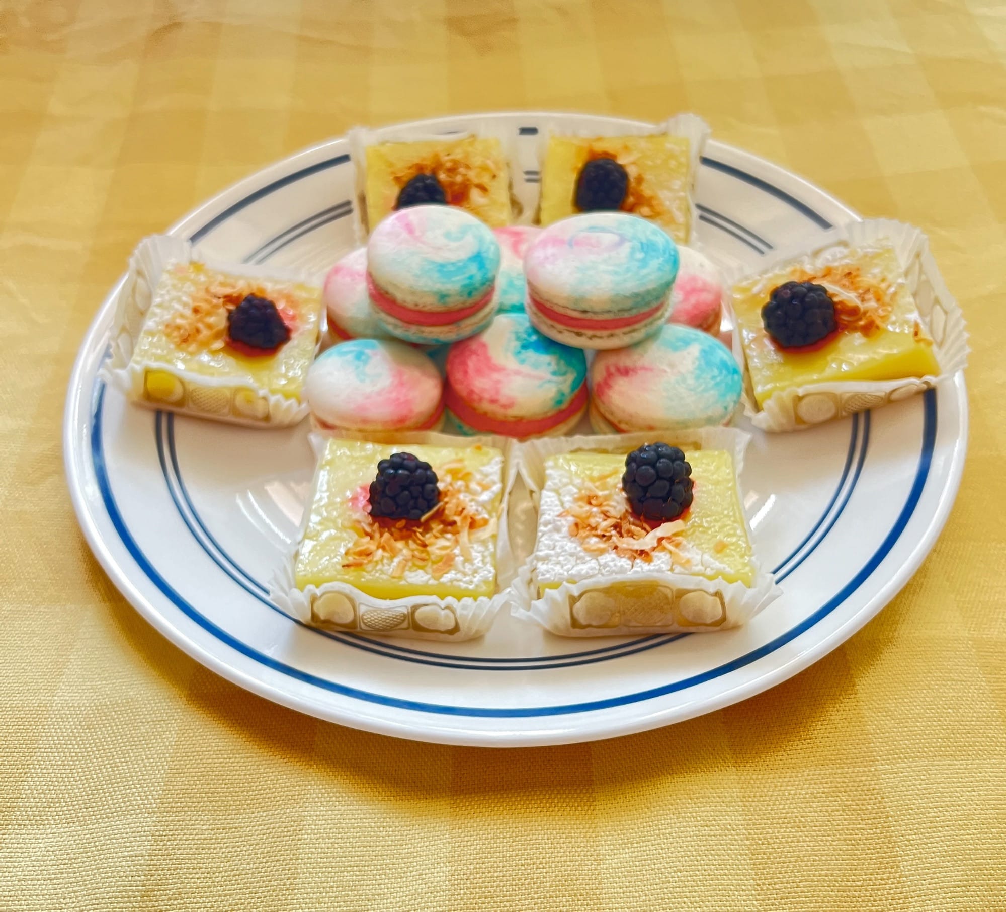 A plate of lemon coconut bars, each with a blackberry on top, under a pile of pink, white, and blue macarons