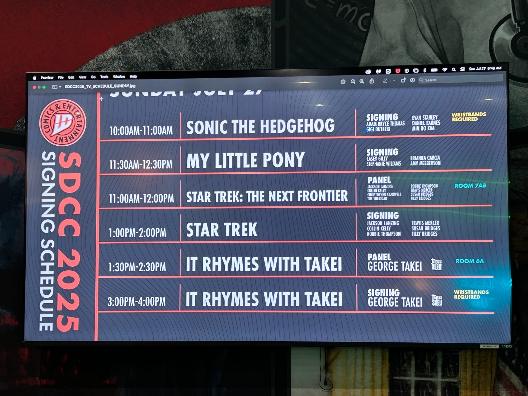 The SDCC 2025 IDW booth signing schedule for Sunday July 27 listed on a television, showing us signing with the rest of the attending Trek comic writers from 1-2 pm (right before George Takei!)
