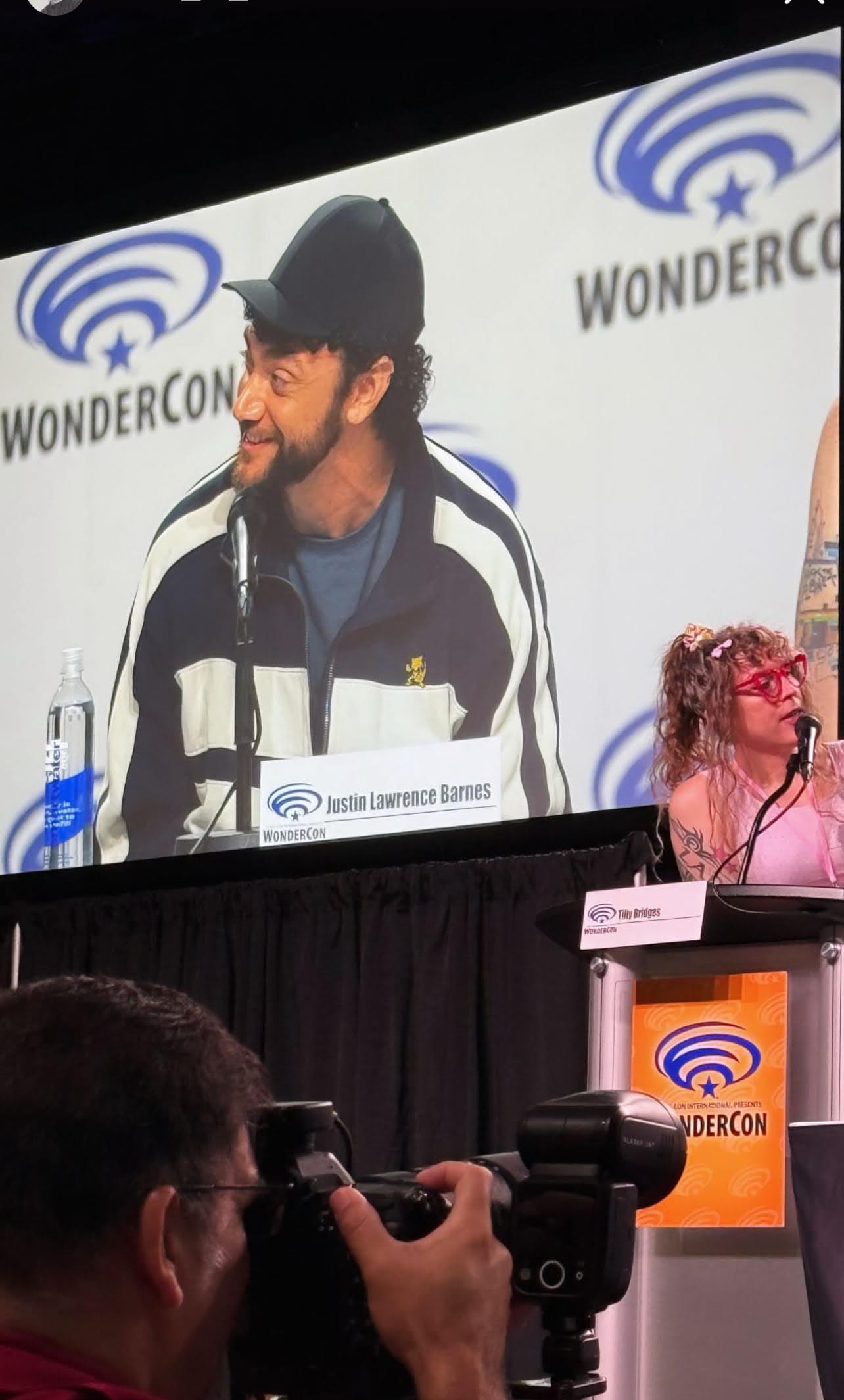 A photographer in the foreground takes photos during the panel, as Tilly is at a the podium moderating and Justin Lawrence Barnes is smiling on one of the video screens behind her