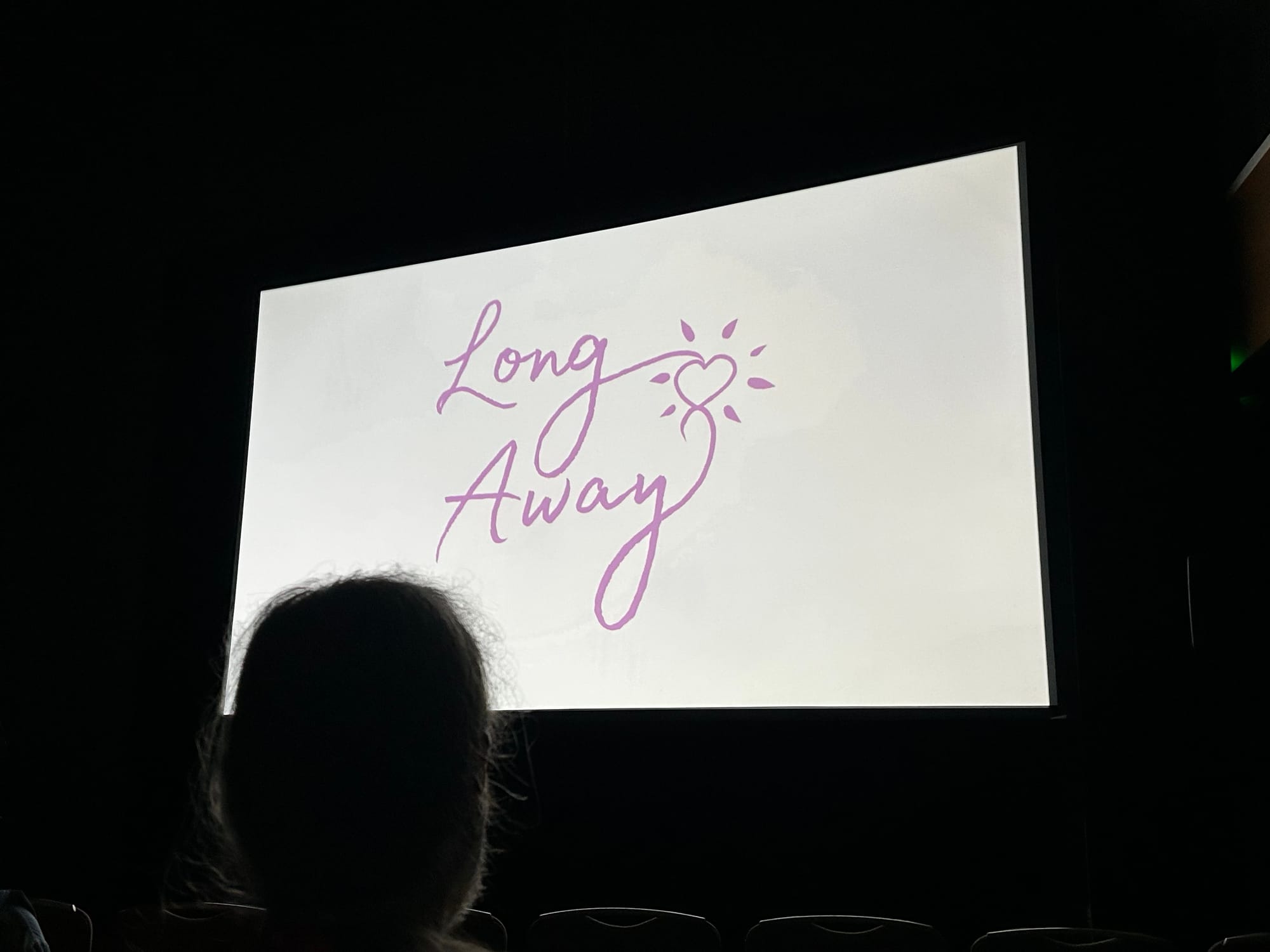 the Long Away title card on a big screen, in a dark room, with a person’s head silhouetted in front of it