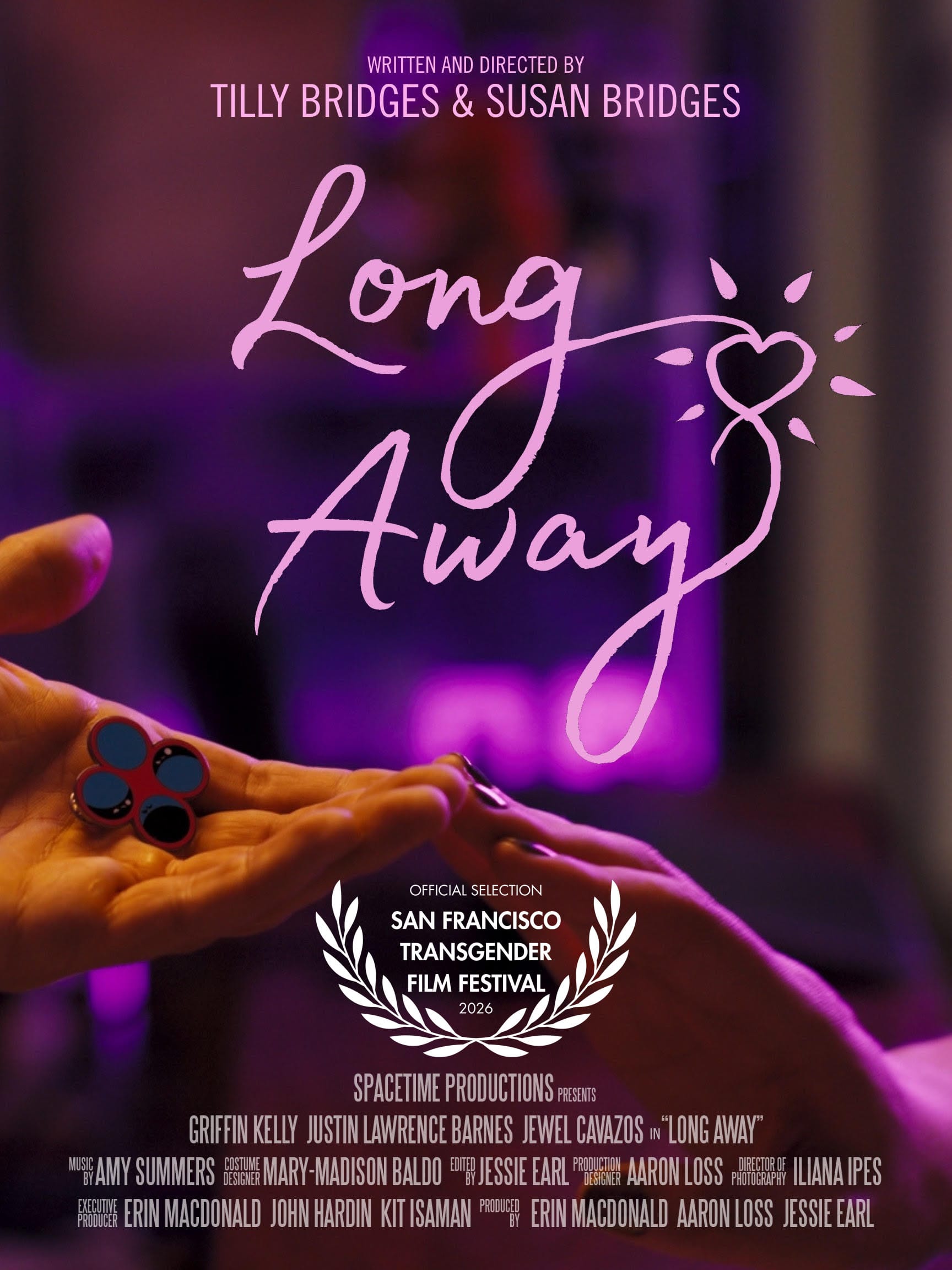 one hand reaching out, palm upward, holding an enamel pin displaying four circles, and another hand reaching out, palm downward, the two hands touch at the fingertips and are bathed in pink light. text reads: LONG AWAY WRITTEN AND DIRECTED BY TILLY BRIDGES & SUSAN BRIDGES SPACETIME PRODUCTIONS PRESENTS GRIFFIN KELLY JUSTIN LAWRENCE BARNES JEWEL CAVAZOS in “LONG AWAY” MUSIC BY AMY SUMMERS COSTUME DESIGN MARY-MADISON BALDO EDITED BY JESSIE EARL PRODUCTION DESIGNER AARON LOSS DIRECTOR OF PHOTOGRAPHY ILIANA IPES EXECUTIVE PRODUCER ERIN MACDONALD JOHN HARDIN KIT ISAMAN PRODUCED BY ERIN MACDONALD AARON LOSS JESSIE EARL Official Selection San Francisco Transgender Film Festival 2026