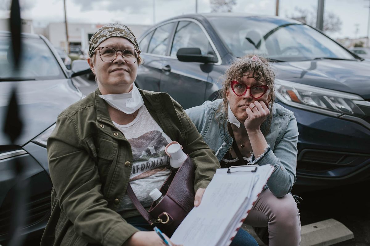 Tilly and Susan sitting down in a parking lot, looking up at the camera monitor during filming on “Long Away”