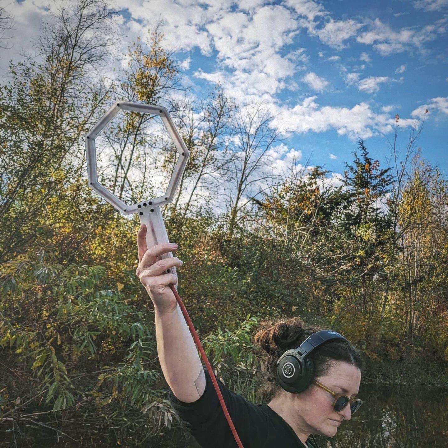 A person with brown hair and sunglasses looks away from the camera and holds up a hexangonal antenna