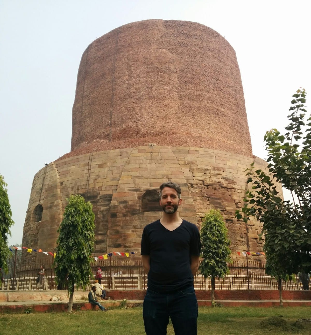 The author standing in front of the Dhamek Stupa, a large, ancient, roughly cylindrical structure of stone and brick.
