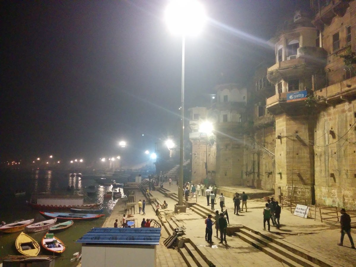 The ghats (stone platforms) alongside the Ganges River in Varanasi, at night, lit by large bright street lights on tall posts, backed by old brick brick or stone buildings, with a loose crowd of people walking, and a dozen or so unoccupied small boats in the river.