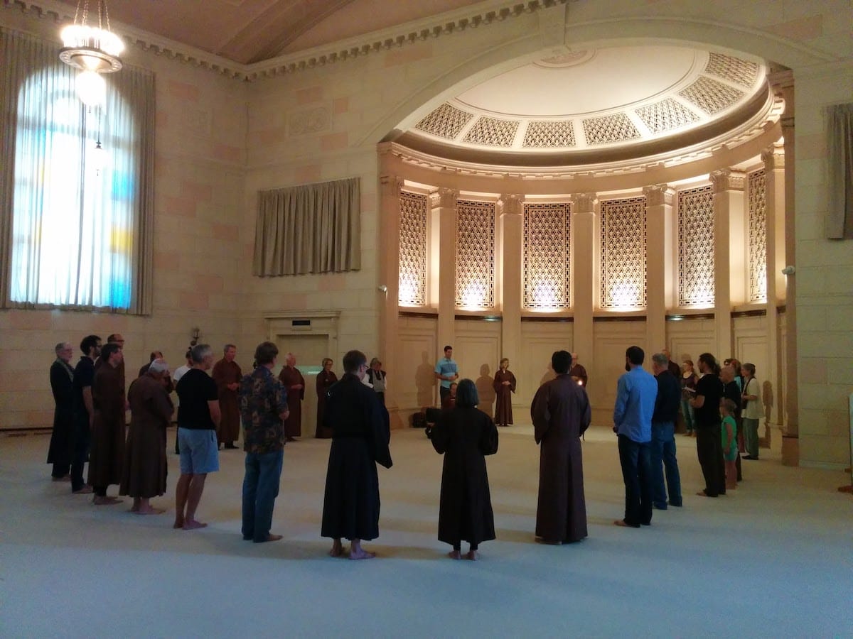 A church sanctuary with very high ceiling, with twenty or thirty people standing in a circle, some wearing Zen robes.