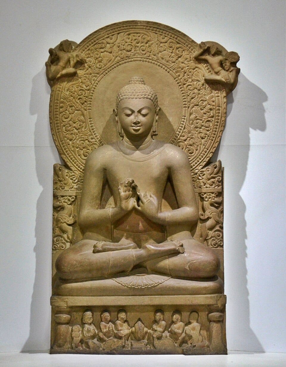 An ancient Indian sandstone sculpture of the Buddha with hands raised in the "preaching the Dharma" mudra.