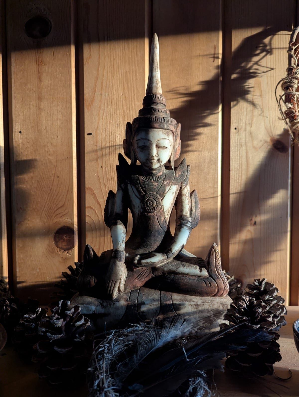 A carved figure of the Buddha in the Earth Witness posture, with one hand touching the ground, surrounded by pine cones and a small bird's nest, against a wall of wood paneling. 
