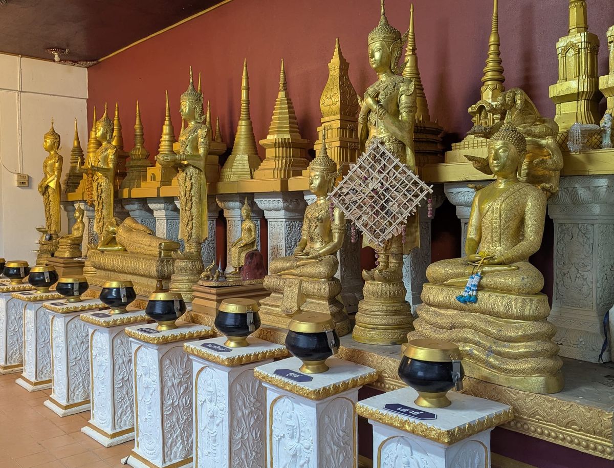 A room with many golden Buddha figures in various postures, with offering bowls on white pillars in front of them, and miniature golden stupas behind.