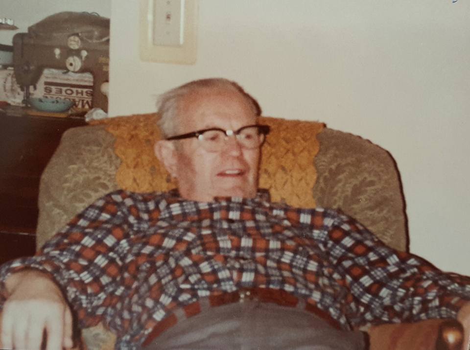 Older man relaxing in an armchair wearing a plaid shirt and glasses, a vintage photograph from the mid-twentieth century.