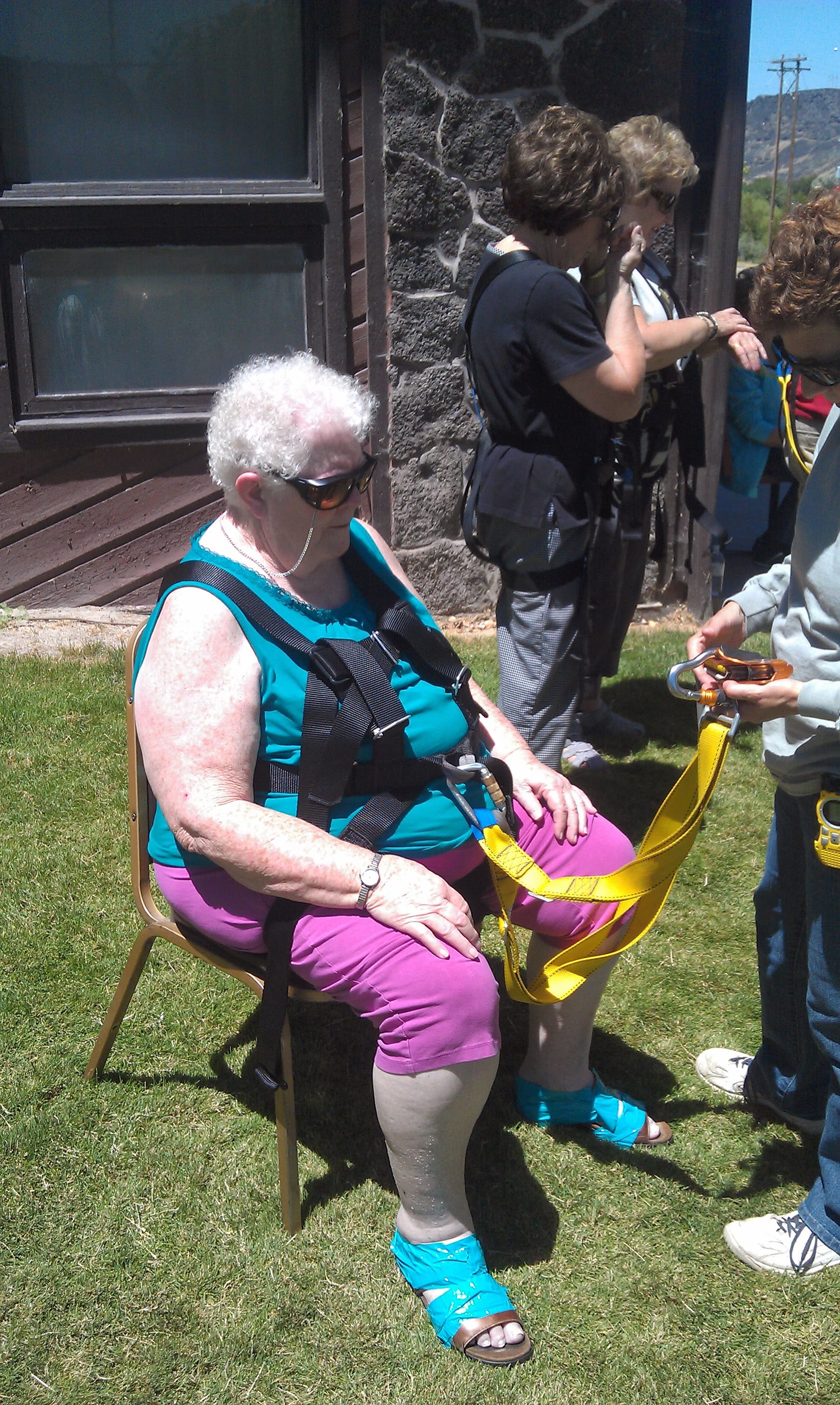 Older woman seated in a harness being fitted by zip line staff outdoors, wearing sandals taped to her feet.