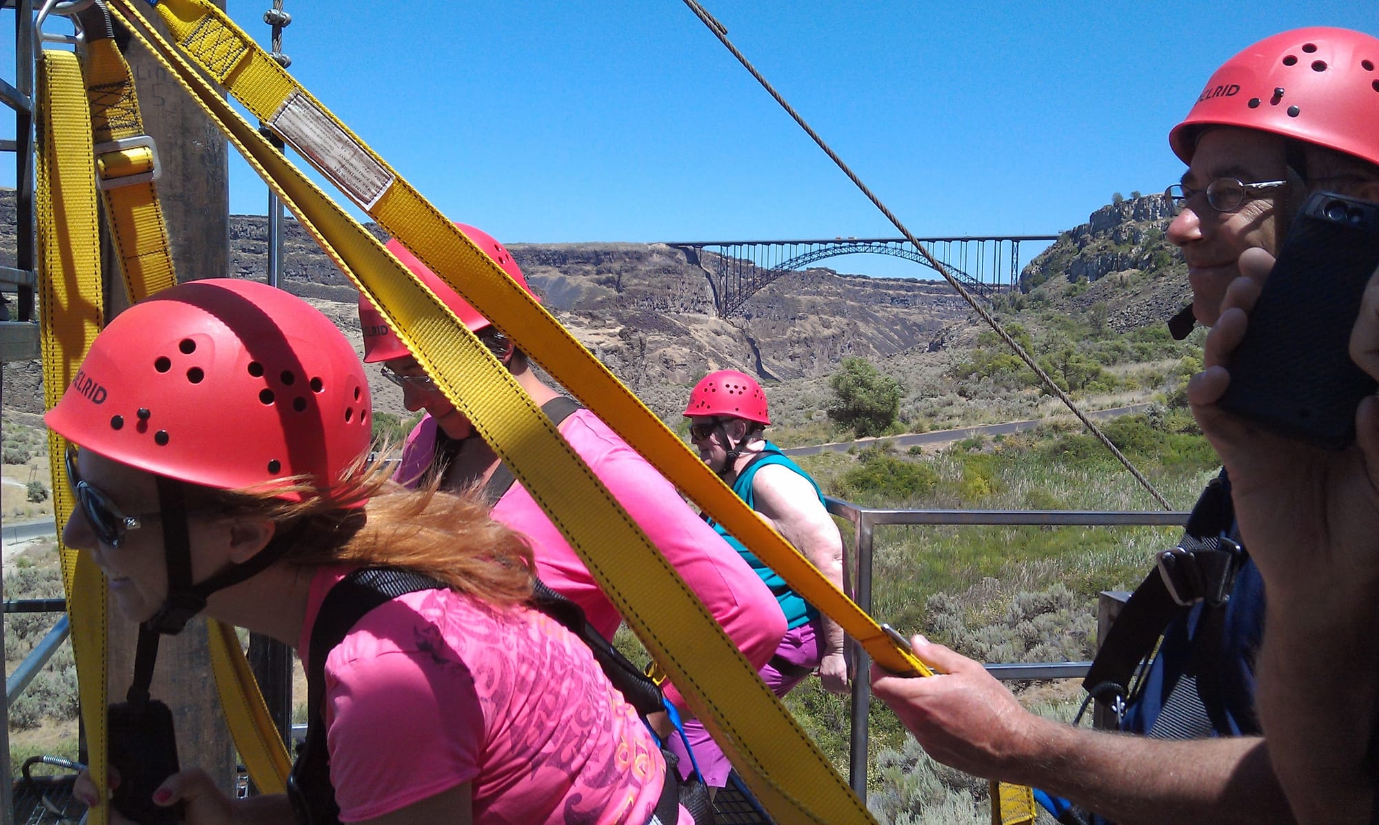 Group of zip line participants in red helmets on a platform with Snake River Canyon and bridge visible behind them.