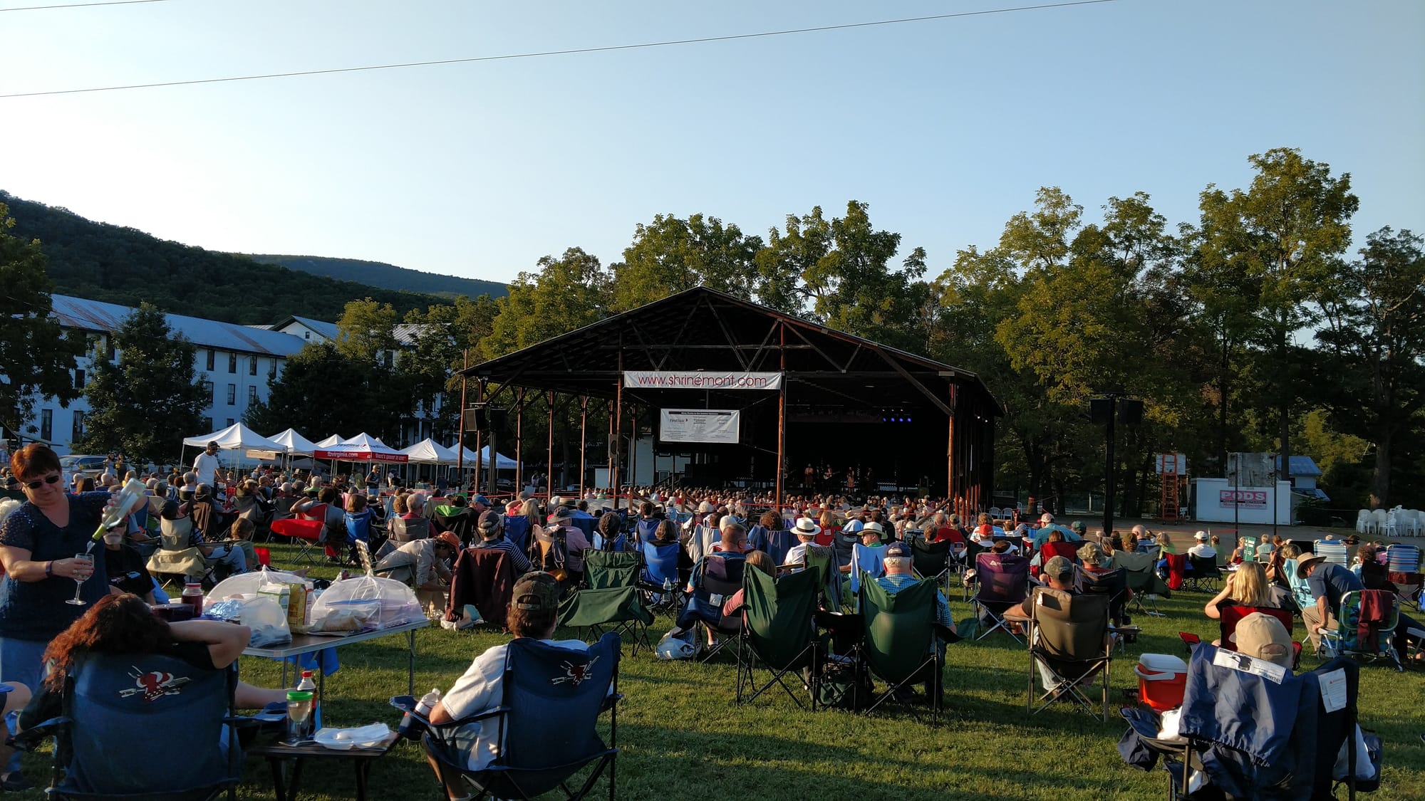 Outdoor music festival crowd seated on grass facing a covered stage at Shrinemont Virginia, surrounded by trees and mountains at golden hour.