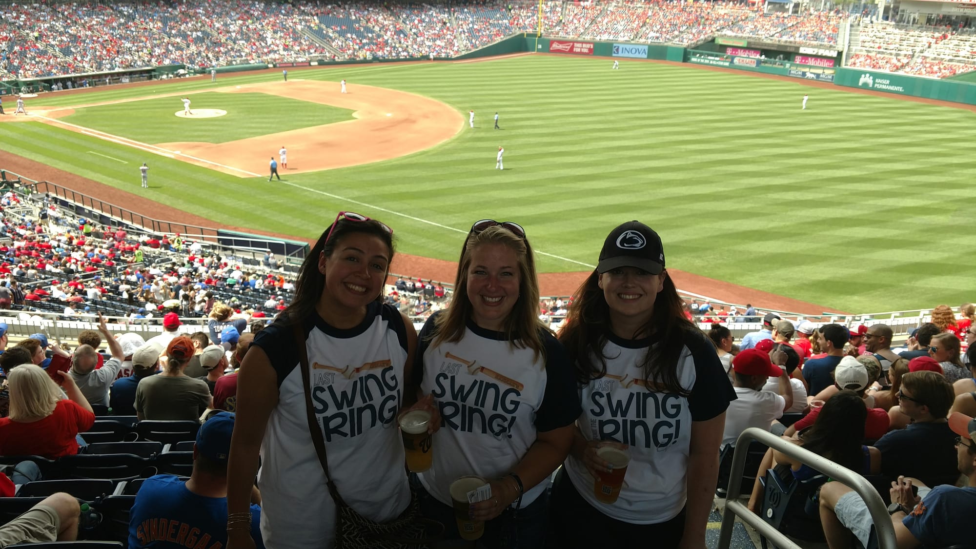Three young women in matching Last Swing Ring shirts smiling in stadium stands with the Nationals baseball field visible behind them.