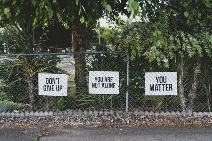 Signs on a fence read “Don’t give up,” “You are not alone,” and “You matter,” surrounded by trees and greenery, conveying hope, support, and encouragement for those struggling.