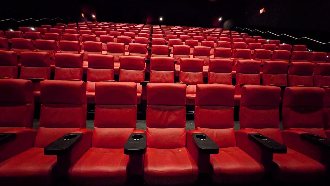 Rows of empty red seats in a dimly lit theater facing a stage with closed red curtains.