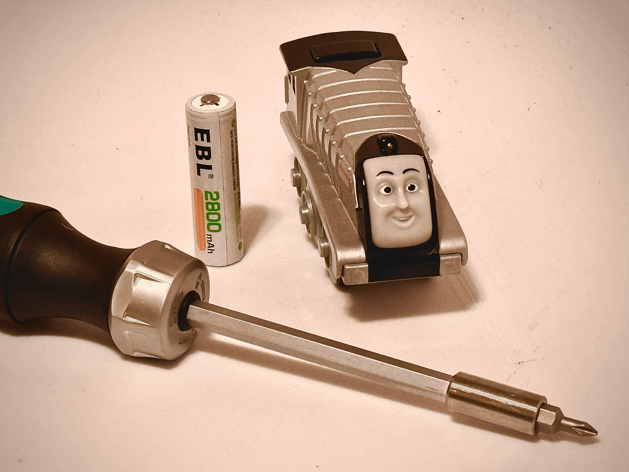 Toy train with a smiling face, a white EBL 2800 mAh AA battery, and a Phillips screwdriver arranged on a light surface. The train is silver with six wheels.