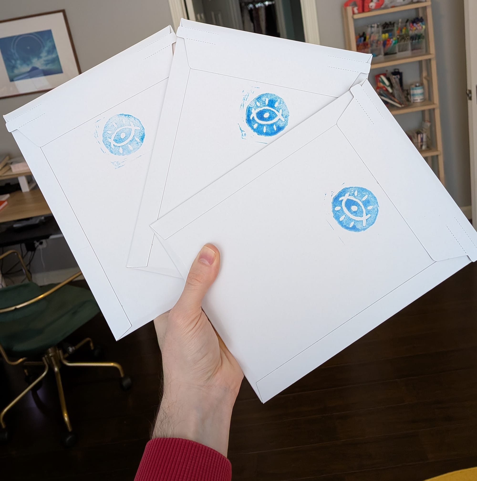A hand holds three cardboard envelops splayed out, with a light blue stamp of a stylized eye on each of them.