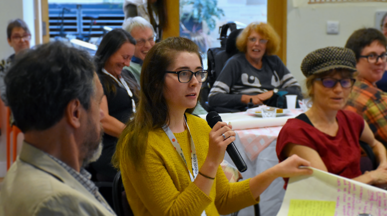A photo of Molly speaking into a microphone, sitting at a table with a large sheet of writing