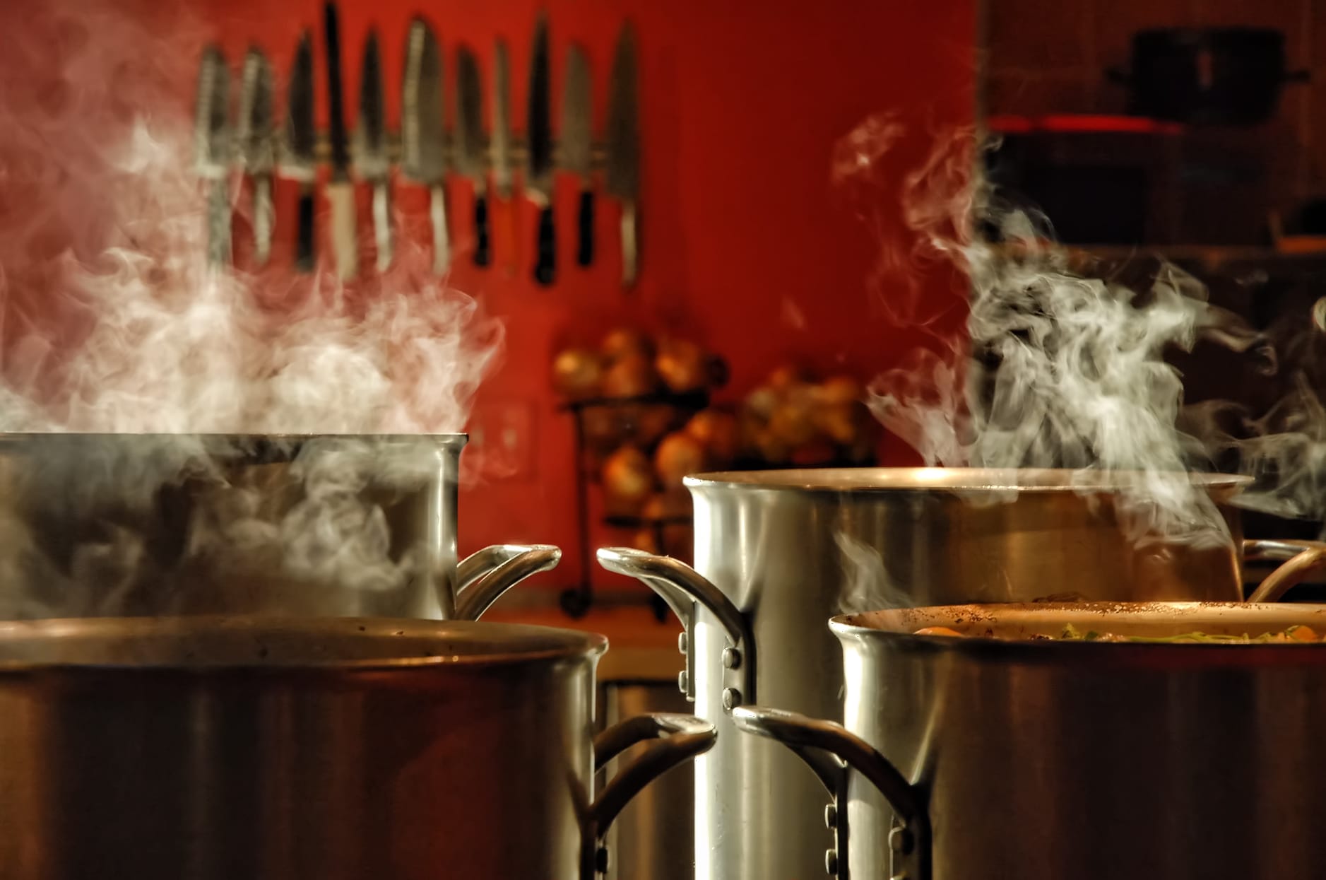 A view of a commercial kitchen with 4 massive stock pots in the foreground and chefs knives on a magnetic rack on the wall in the background.