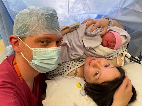 Me, a woman with dark hair, lying on a bed holding our new baby, with my husband, a man wearing hospital scrubs