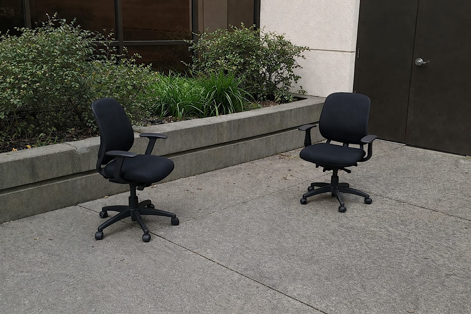 Two office chairs left outside near a planter and plain building wall.