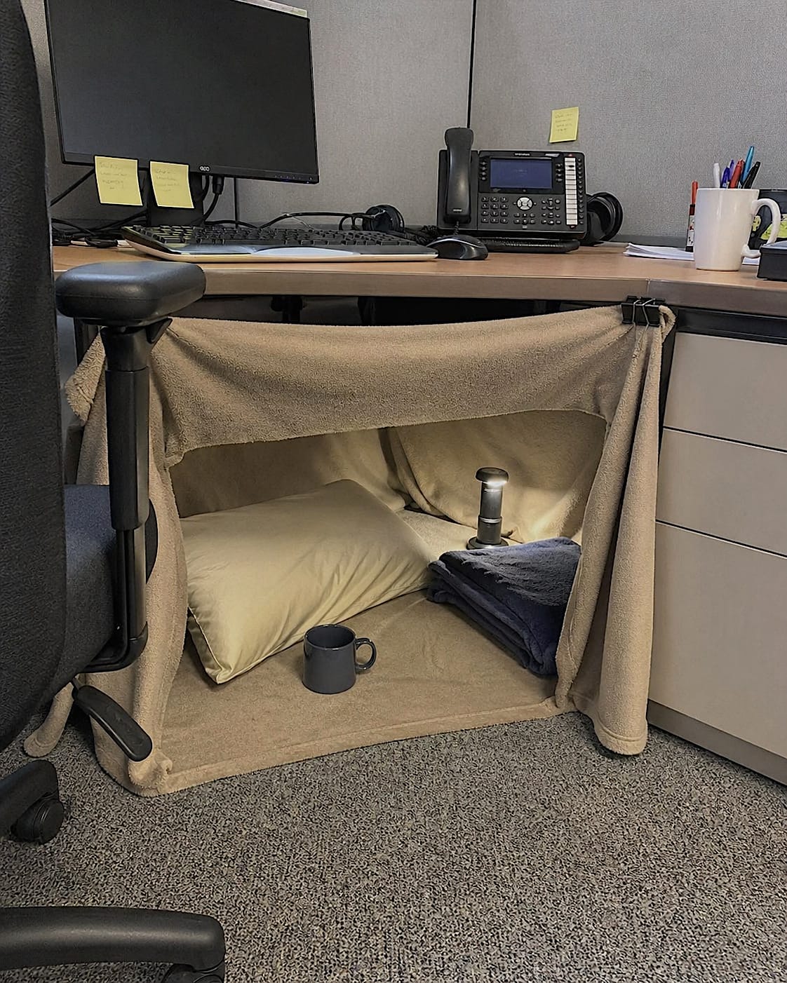 Office cubicle with a small blanket fort, pillow, mug, and lamp tucked under the desk.