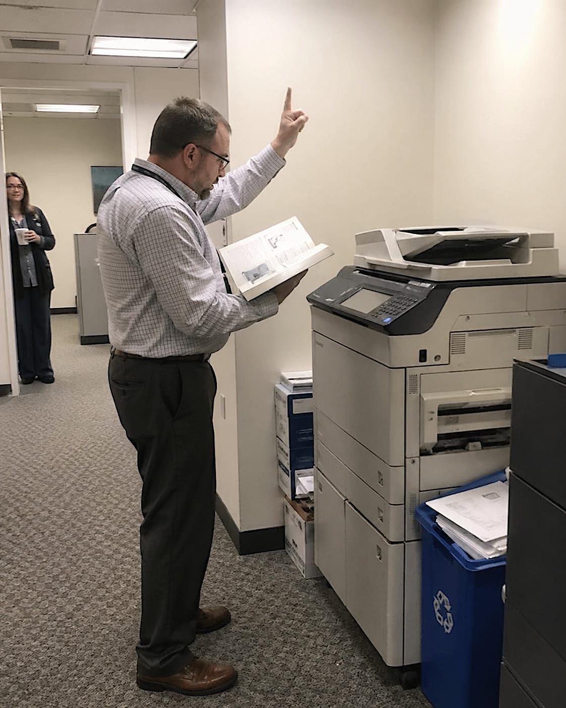 Office worker holding an open printer manual and gesturing dramatically toward a large office printer.