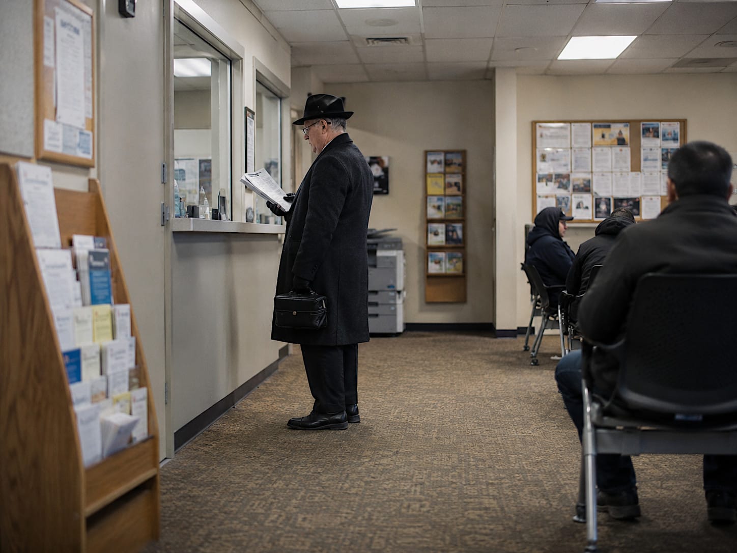Older man in a dark overcoat and brimmed hat standing in a plain municipal-building hallway.