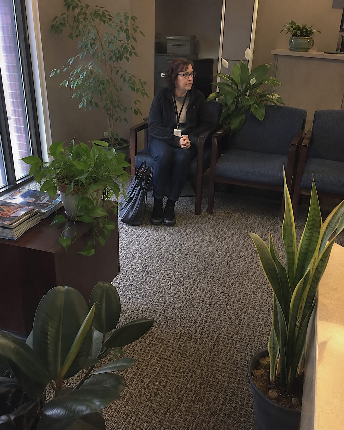 An adult visitor sits alone in a waiting chair in a bland office reception area, with potted plants placed around the room so they visually surround her.