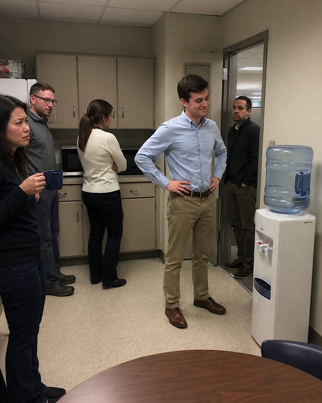 Young intern standing proudly beside an office water cooler while coworkers react with concern.