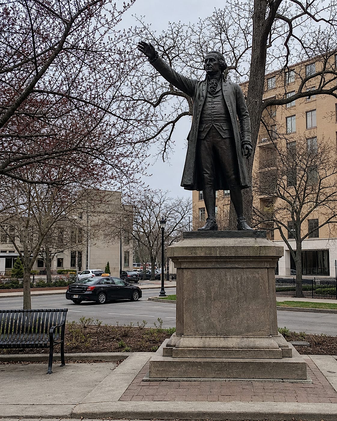 Bronze statue in a city plaza with its raised arm lined up with a passing taxi.