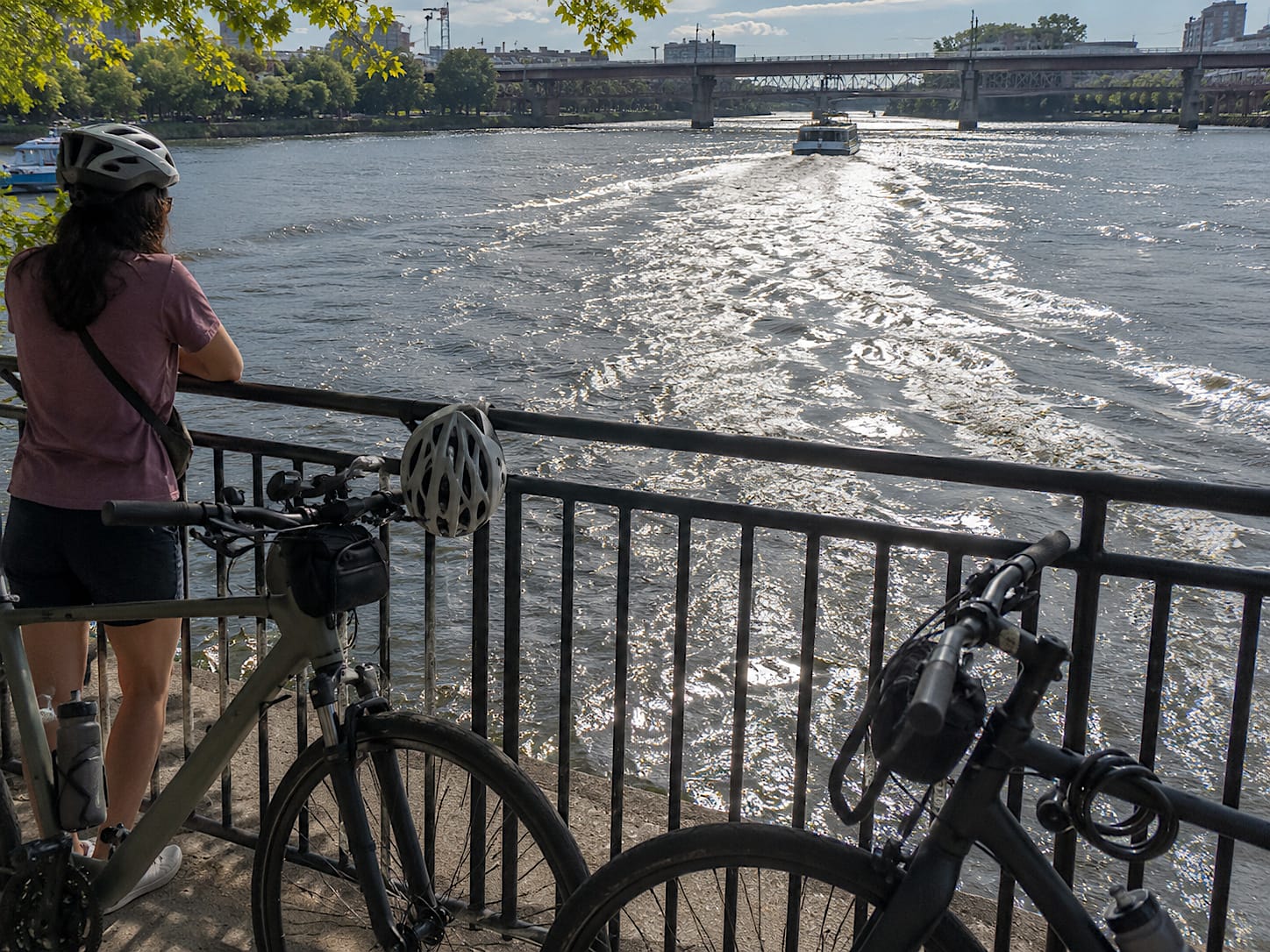 Bikes and helmets by a riverside railing as a boat wake breaks sunlight into bright zigzags on the water.