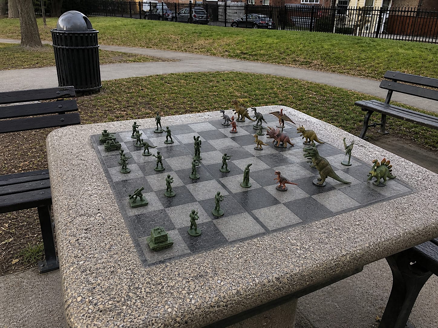 Outdoor chess table in a park with toy dinosaurs facing green plastic army men instead of chess pieces.