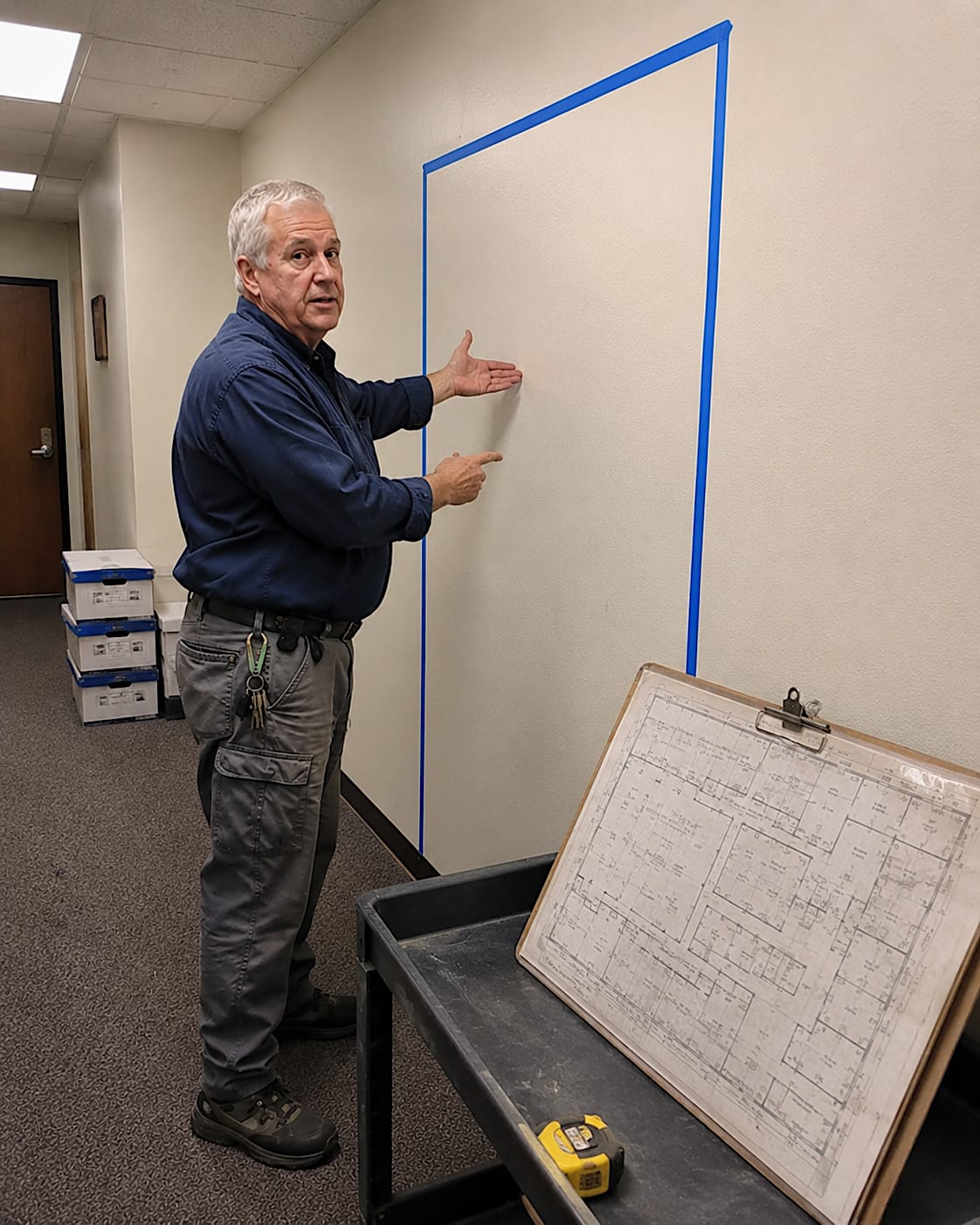 Maintenance man pointing to a taped door outline on a blank office corridor wall beside an old floor plan.