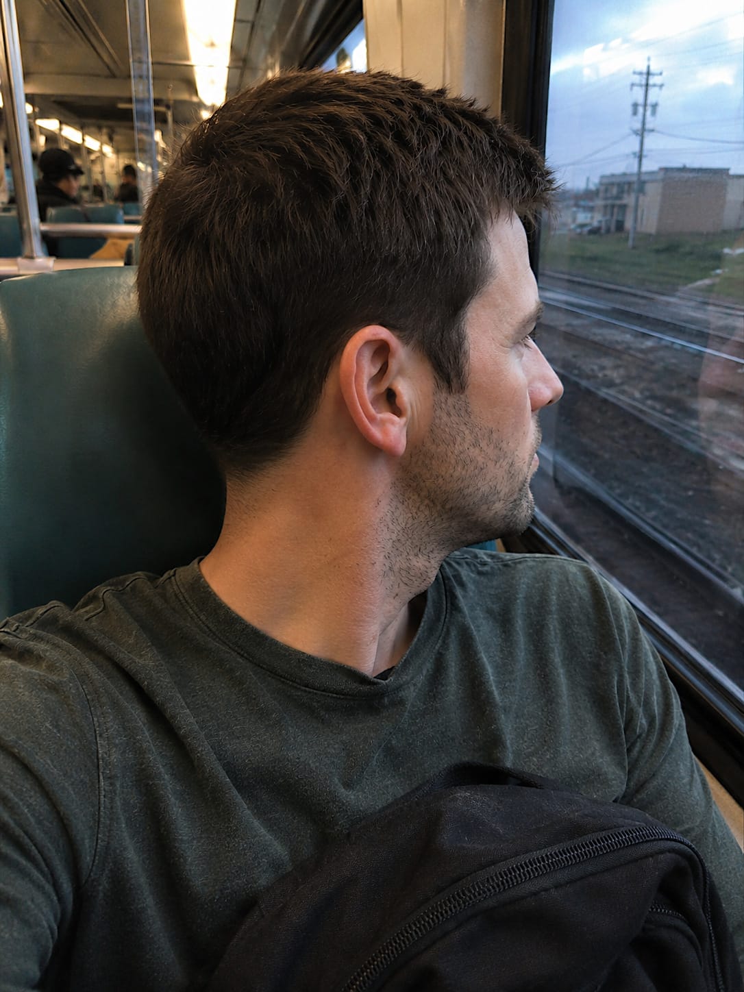 Close train selfie of Evan leaning back, turned toward the window, with his ear and partial profile in front as tracks pass in early evening light.