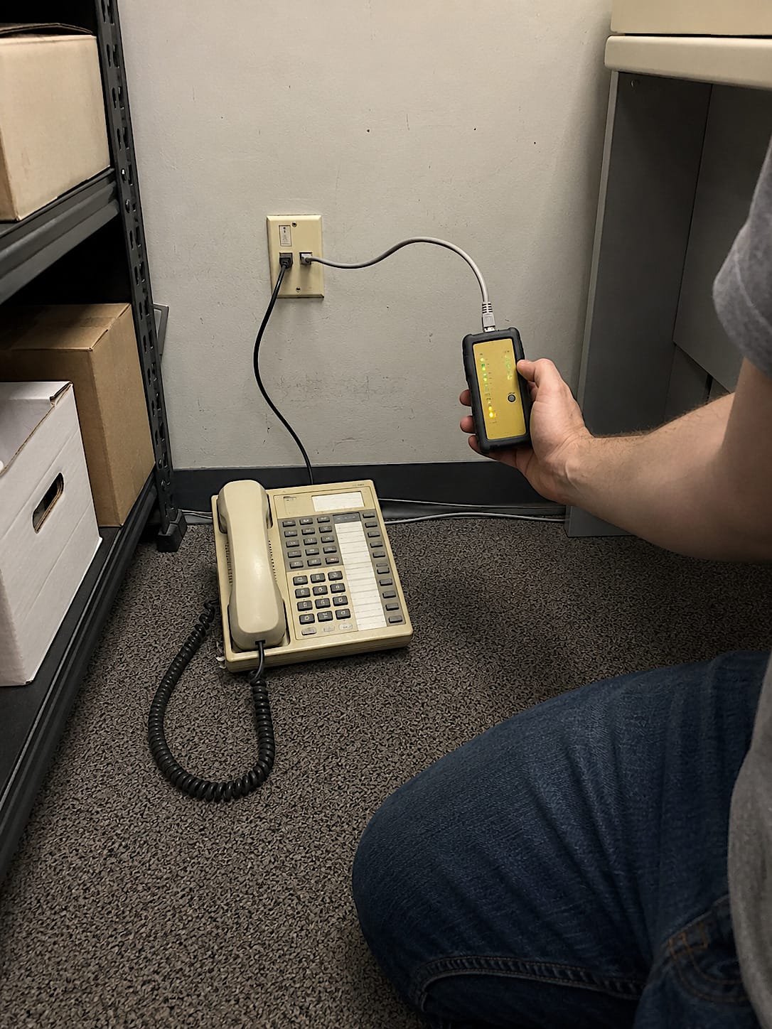Person using a cable tester on an old office wall jack connected to a retired beige desk phone.