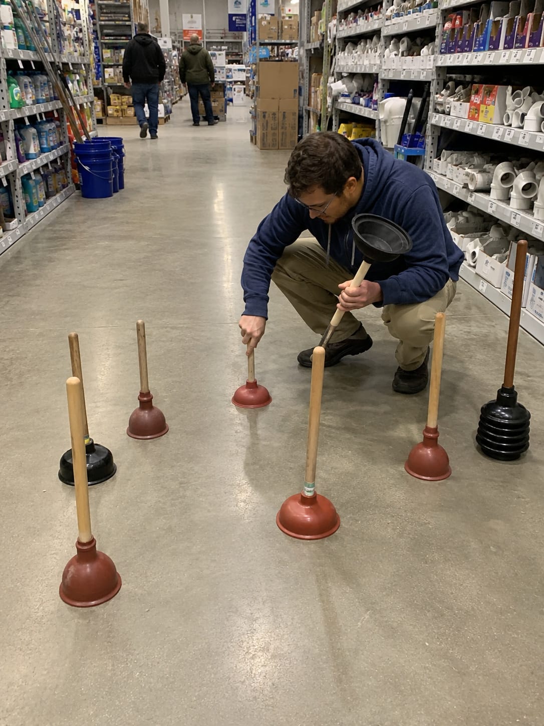 Customer in a hardware-store plumbing aisle tests plungers stuck to the floor while other plungers wait nearby.