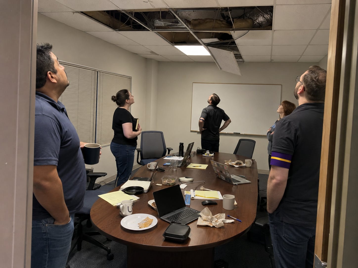 Coworkers stare up at open ceiling tiles above a cluttered conference table after a meeting appears to have vanished.E