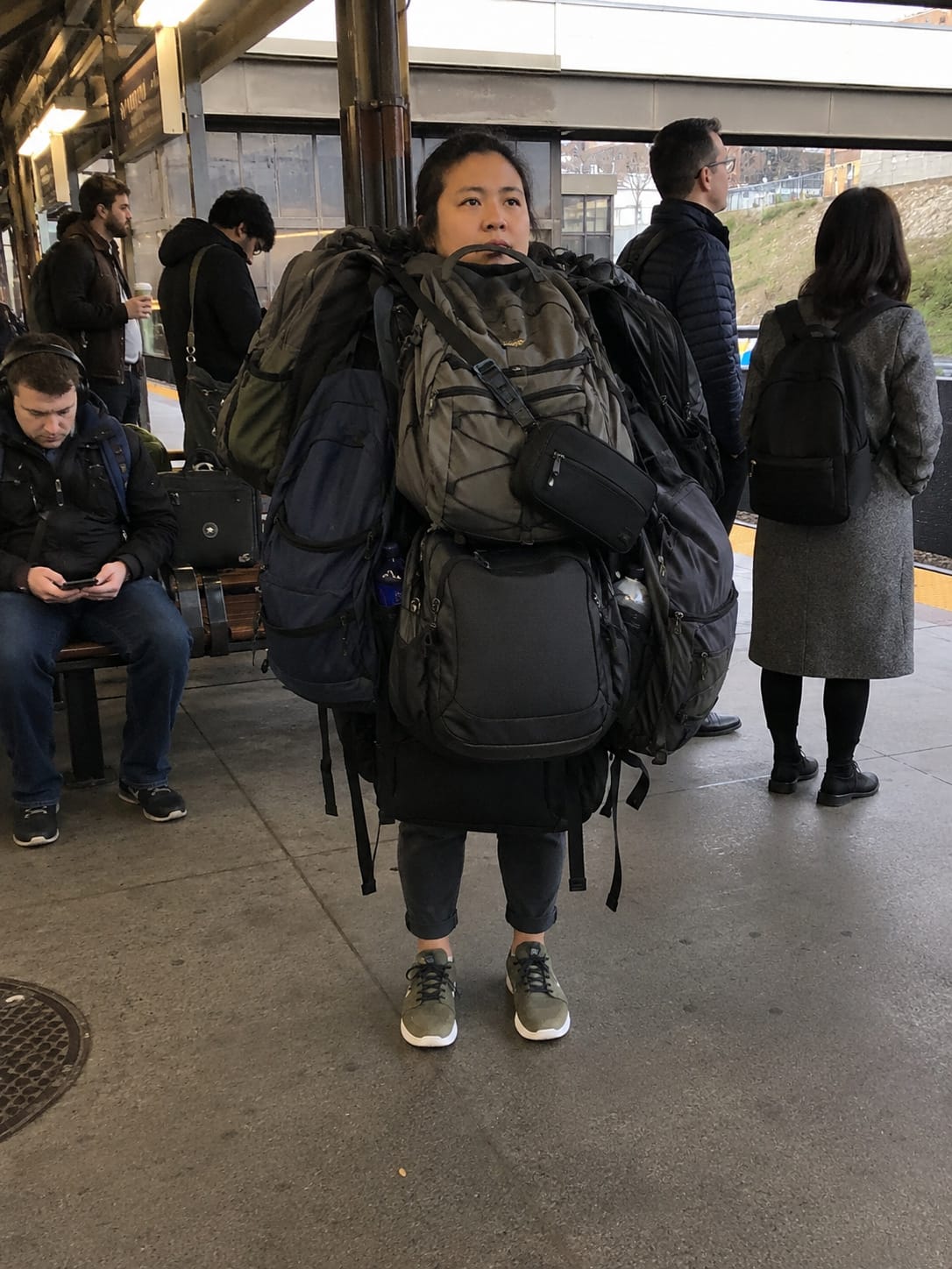 A commuter waits on a train platform covered in backpacks, as if the bags packed one human for emergencies.