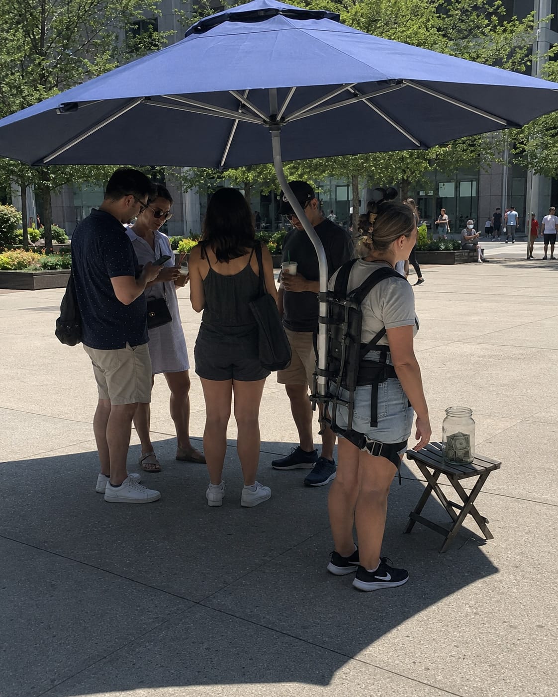Woman with a patio umbrella strapped to her back shades several people in a sunny plaza, with a tip jar nearby.